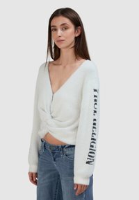 Weißer, weicher, strukturierten Pulli mit tiefem V-Ausschnitt und verdrehter Front. Schwarzer Text auf dem linken Ärmel steht im Kontrast zu dem hellen Stoff.