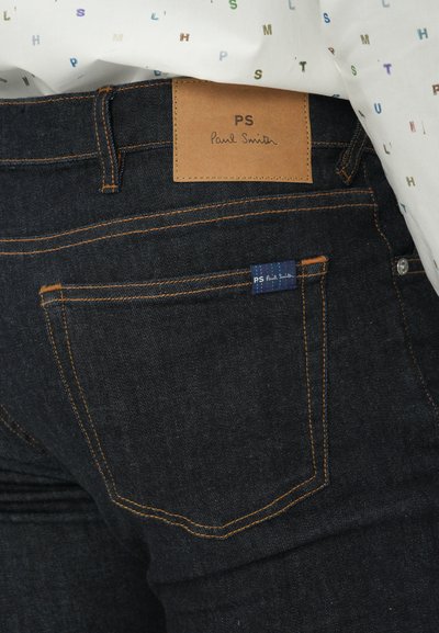 Jean en denim foncé avec une patte en cuir marron étiquetée "PS Paul Smith", des coutures orange et une petite étiquette bleue sur la poche arrière.