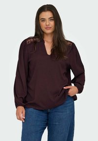 Blouse bordeaux à manches longues avec un décolleté en V, en tissu texturé, avec des accents en dentelle sur les épaules et une coupe décontractée. Associée à un jean bleu.