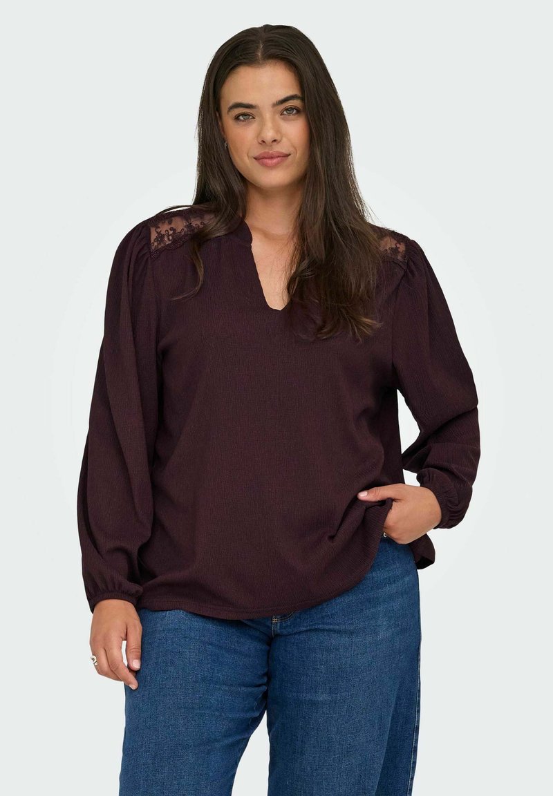 Blouse bordeaux à manches longues avec un décolleté en V, en tissu texturé, avec des accents en dentelle sur les épaules et une coupe décontractée. Associée à un jean bleu.