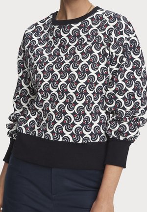 Mikina s černobílým spirálovým vzorem a malými červenými tečkami, doplněná tmavými kalhotami a zlatým náhrdelníkem, zobrazena od hrudi po pas.