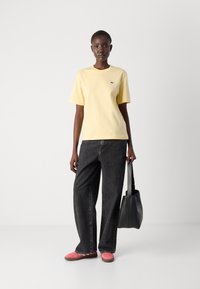 Gul bomull t-skjorte med korte ermer og en liten logo, kombinert med løse svarte jeans og rosa sko. Holder en svart tote-veske.