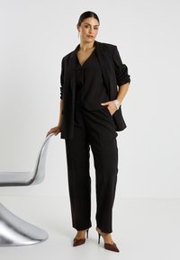Traje negro a medida con blazer entallado y pantalones de pierna ancha. Tacones marrones brillantes y accesorios plateados complementan el conjunto. Textura suave.