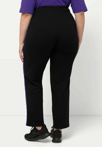 Zwarte sportbroek met een recht model, voorzien van paarse zijstrepen en een elastische tailleband. Gekleed met zwarte sneakers.