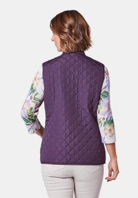 Gep quilting-Weste in tiefem Lila mit hohem Kragen. Sie hat eine glatte Textur und diagonale Nähte, kombiniert mit einem floralen Langarmshirt darunter.
