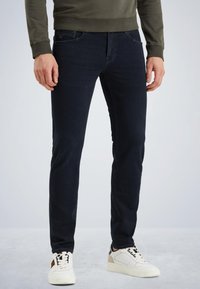 Jeans slim fit bleu foncé en denim, avec des poches avant et un logo subtil. Associés à des baskets blanches et un sweat-shirt vert olive.