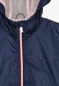 Giacca a vento blu navy con cappuccio foderato in rete, zip bianca con strisce rosse e argento, dotata di un piccolo logo vicino all'orlo.