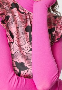 Růžový dlouhý rukáv top s květinovým vzorem na panelu, který má volánkové detaily. Měkká textura; úzké rukávy; výrazný černý květinový design na světle růžovém pozadí.