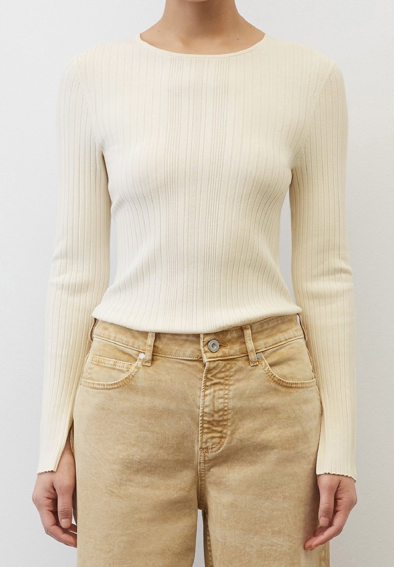 Personne portant un pull crème à manches longues en côtes, rentré dans un jean marron clair taille haute, debout devant un fond blanc uni.