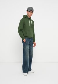 Zaļš kapucis ar priekšējo kabatu un baltām auklām, saskaņots ar brīviem ziliem džinsiem ar iešūtām rakstzīmēm, un baltām sporta apavām.