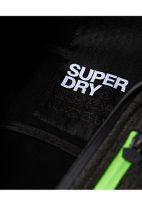 Étiquette intérieure noire avec "SUPER DRY" en lettres blanches, entourée d'un tissu noir texturé avec un accent de zip vert.