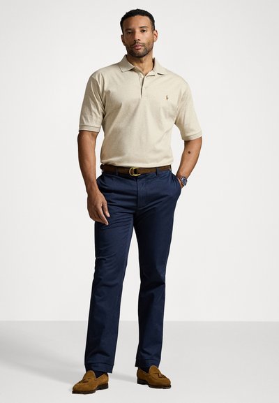 Cămașă polo bej, pantaloni bleumarin, curea maro și pantofi maro din daim. Cămașa polo are guler și mâneci scurte; pantalonii sunt drepți.