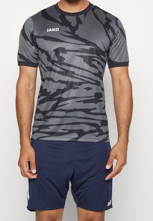 Homme portant un maillot de sport JAKO gris et noir à motif abstrait et un short bleu marine avec détails blancs, debout sur un fond uni.