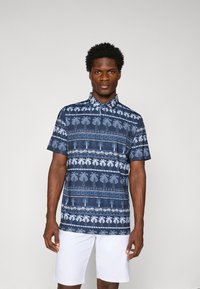 Polo a maniche corte blu navy con motivo a palme, colletto con bottoni e vestibilità rilassata, abbinato a pantaloni corti bianchi.