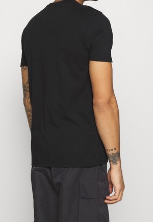 Mężczyzna w prostym czarnym t-shircie z krótkim rękawem i ciemnych spodniach typu cargo, pokazujący plecy i lewą rękę z tatuażami motyla i tekstu.