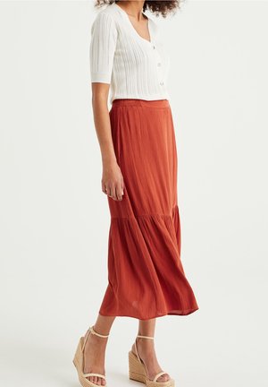 Vrouw draagt een wit kortmouwig shirt met knopen, een roestkleurige lange rok en beige sleehaksandalen, staand tegen een effen achtergrond.