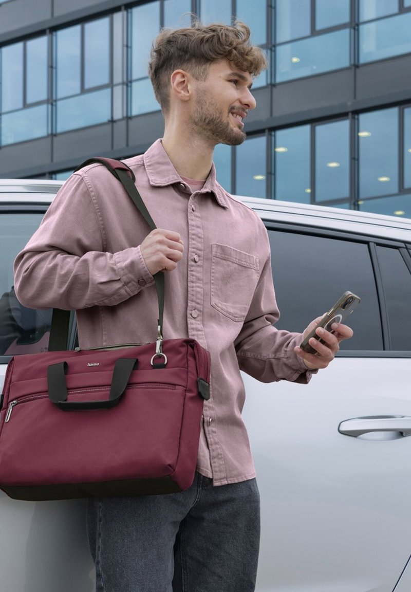 Junger Mann in einem rosa Hemd, der eine kastanienbraune Umhängetasche trägt, ein Smartphone hält und neben einem weißen Auto vor einem gläsernen Gebäude steht.