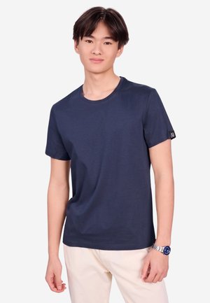 Junger Mann trägt ein marineblaues Kurzarm-T-Shirt, helle Hosen und eine Armbanduhr, steht vor einem weißen Hintergrund.