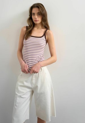 Young woman with long hair wearing a brown-striped sleeveless top and white wide-leg shorts standing against a plain background.