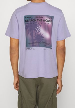 Mężczyzna w lawendowym t-shircie z nadrukiem palm i tekstem "Search the World" na plecach, zestawiony z zielonymi spodenkami typu cargo.