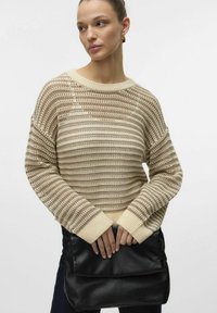 Gebreide trui in beige met gestreept patroon en ribbelzoom. Model houdt een zwarte leren clutch vast. Zachte textuur, ontspannen pasvorm, brede mouwen.