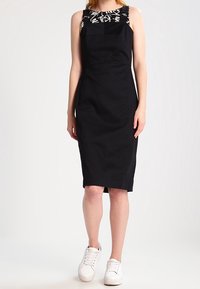 Kobieta stoi w czarnej sukience bez rękawów, z białym wzorzystym topem pod spodem i białych trampkach, na jednolitym tle.