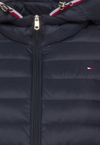Veste matelassée bleu marine avec fermeture éclair centrale dorée, cordons rayés rouge-blanc-bleu et un petit logo drapeau rouge-blanc-bleu sur la poitrine.
