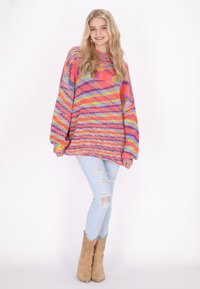 Pulover oversize tricotat multicolor, cu guler rabatabil, mâneci lungi și o croială lejeră, asortat cu blugi deschiși la culoare, uzati, și cizme maro.