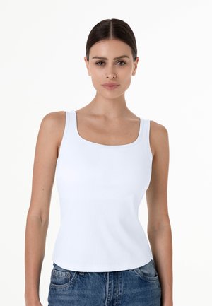 Donna con capelli scuri legati indietro che indossa una canottiera bianca a coste e jeans blu, rivolta in avanti su uno sfondo bianco semplice.