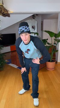 Zwarte beanie met blauwe letters, lichtblauw shirt met lange mouwen, donkere spijkerbroek en witte sneakers. Staande in een kamer met planten en meubels.