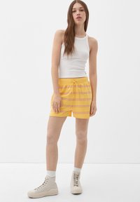 Gelb gestreifte Shorts mit einem Kordelzugbund und Seitentaschen, kombiniert mit einem weißen gerippten Tanktop und beigen High-Top-Schuhen.