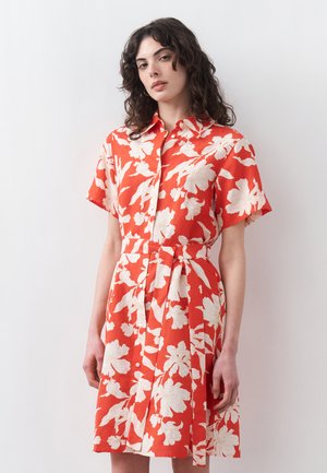 Vrouw met krullend haar, gekleed in een oranje jurk met korte mouwen, grote witte bloemenpatroon en een bijpassende tailleband.