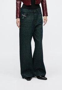 Sötét, széles szárú farmernadrágot viselő személy, enyhén kopottas hatással, fekete csizmákkal, fekete felsővel és rövidített piros bőrkabáttal.