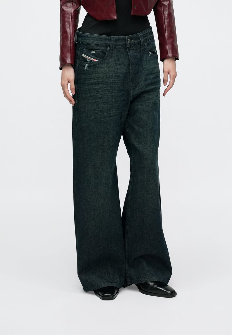 Sötét, széles szárú farmernadrágot viselő személy, enyhén kopottas hatással, fekete csizmákkal, fekete felsővel és rövidített piros bőrkabáttal.