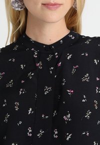 Svart blus med delikata blommönster i rosa och vitt, med mandarinkrage och knäppning fram.