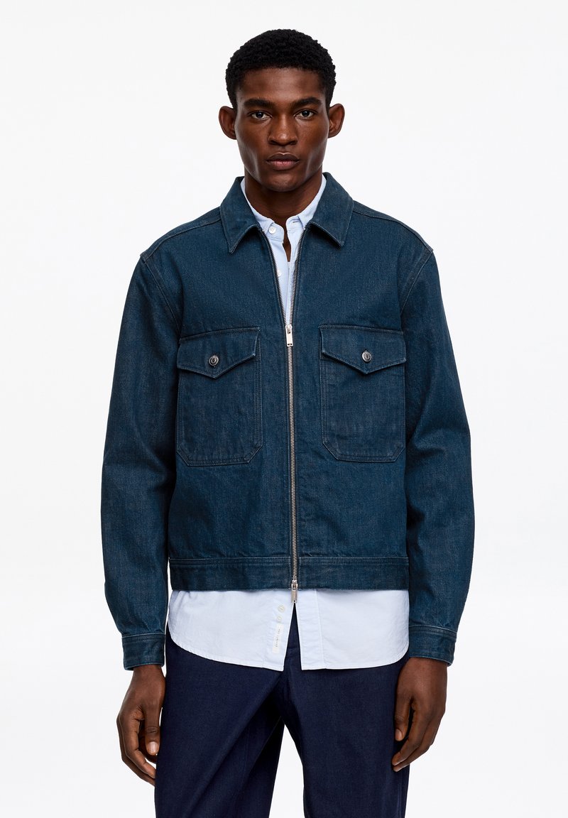 Homme portant une veste en jean bleu foncé à fermeture éclair sur une chemise blanche à boutons et un pantalon bleu marine, debout devant un fond blanc uni.