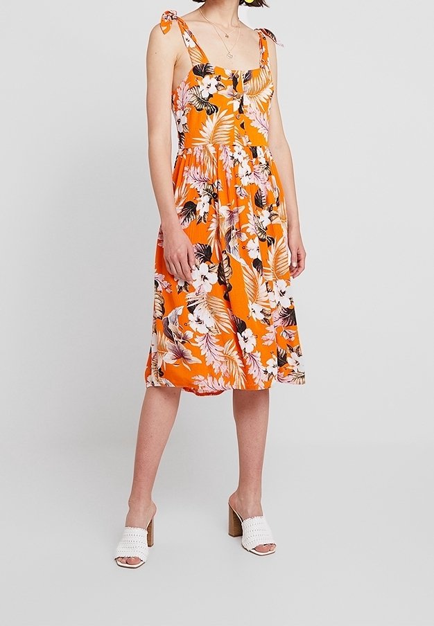 Femme portant une robe d'été orange à fleurs sans manches avec des bretelles à nouer et des sandales blanches à talons, debout devant un fond clair uni.