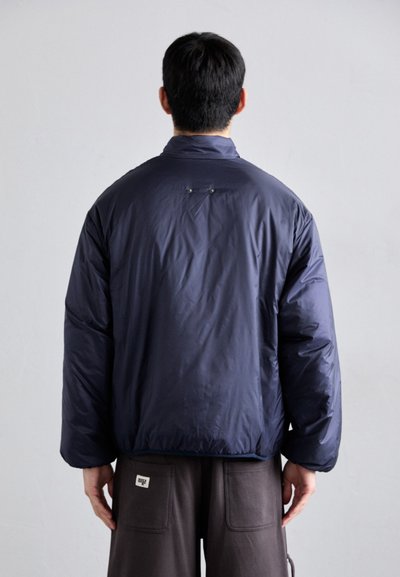 Giacca blu navy imbottita con colletto alto e vestibilità oversize. Tessuto liscio e design minimalista; presenta due piccoli bottoni metallici sul retro.