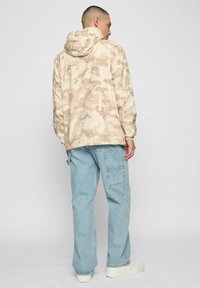 Beżowa kurtka moro z kapturem, lekki materiał, elastyczne mankiety, w zestawie z jasnoniebieskimi jeansami cargo i białymi trampkami.