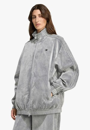 Donna che indossa una giacca sportiva in velour grigia con collo alto, cerniera, tasche laterali e un discreto logo Adidas sul petto.