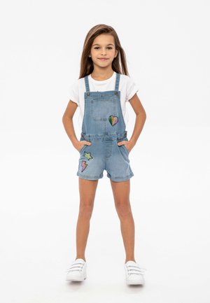 Salopette en denim bleu clair avec des poches à rabat et des designs brodés colorés : un cœur, une étoile et un éclair. T-shirt blanc et baskets.