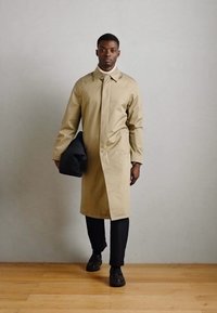 Beige lichtgewicht jas met een rechte snit, lange mouwen en een kraag, gedragen over donkere broeken, gecombineerd met een zwarte tas en bruine schoenen.