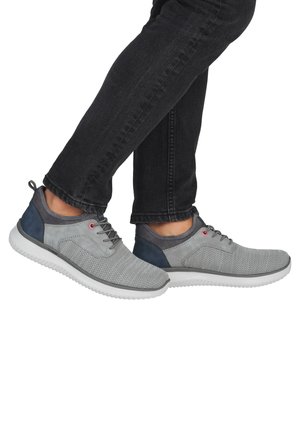 Personne portant des baskets en maille grise avec des accents bleu marine et des semelles blanches, associées à un jean noir, les pieds croisés à la cheville.