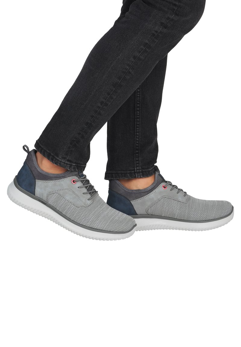 Person trägt graue Stricksneaker mit marineblauen Akzenten und weißen Sohlen, kombiniert mit schwarzen Jeans, die Füße sind an den Knöcheln gekreuzt.