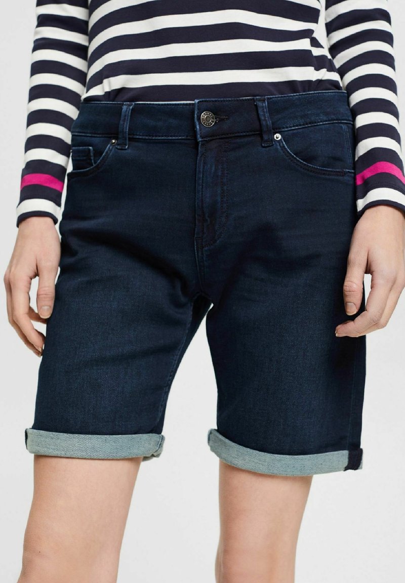 Pantalones cortos de mezclilla azul oscuro con dobladillos enrollados, con cierre de botón, bolsillos delanteros y una textura suave. Diseñados para un ajuste ceñido.