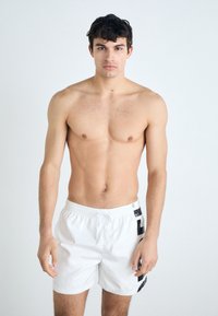 Shorts de bain blancs avec une taille à cordon de serrage et des accents noirs sur les côtés. Le modèle a les cheveux courts et foncés et est torse nu, se tenant devant un fond clair.