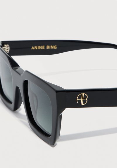 Schwarze rechteckige Sonnenbrille mit dicken Rahmen, grauen Gläsern, goldfarbenem "AB"-Logo am Bügel und "ANINE BING" auf der Innenseite des Bügels gedruckt.