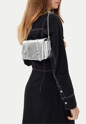 Kobieta ubrana w czarną sukienkę z długim rękawem z białymi przeszyciami, z metaliczną srebrną torebką na ramię z łańcuszkowym paskiem na plecach.
