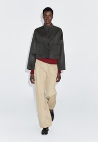 Wysoki mężczyzna w ciemnoszarym zapinanym żakiecie, pod którym ma na sobie czerwoną i czarną pasiastą bluzkę, beżowych szerokich spodniach i czarnych butach, idzie do przodu.