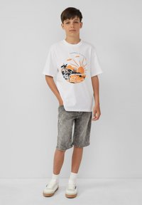 Biała bawełniana koszulka z pomarańczowym i czarnym graficznym wzorem na temat plaży, zestawiona z szarymi dżinsowymi szortami. Sneakersy z białymi i beżowymi akcentami.